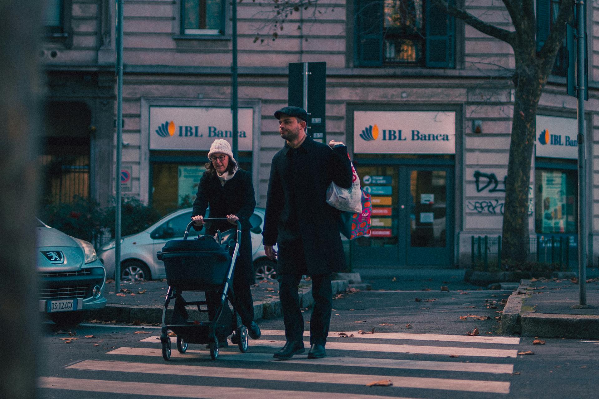 Couple walking with baby stroller across a street in a city setting, near IBL Banca.