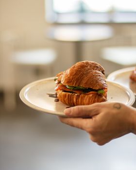 Close-up of a fresh croissant sandwich with salad held by a woman in a cafe setting.