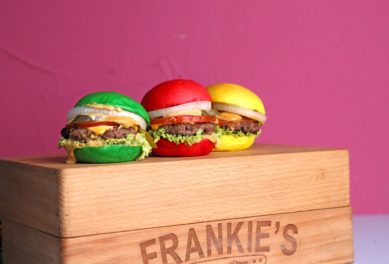three vibrant burgers with colored buns on a wooden box against a pink