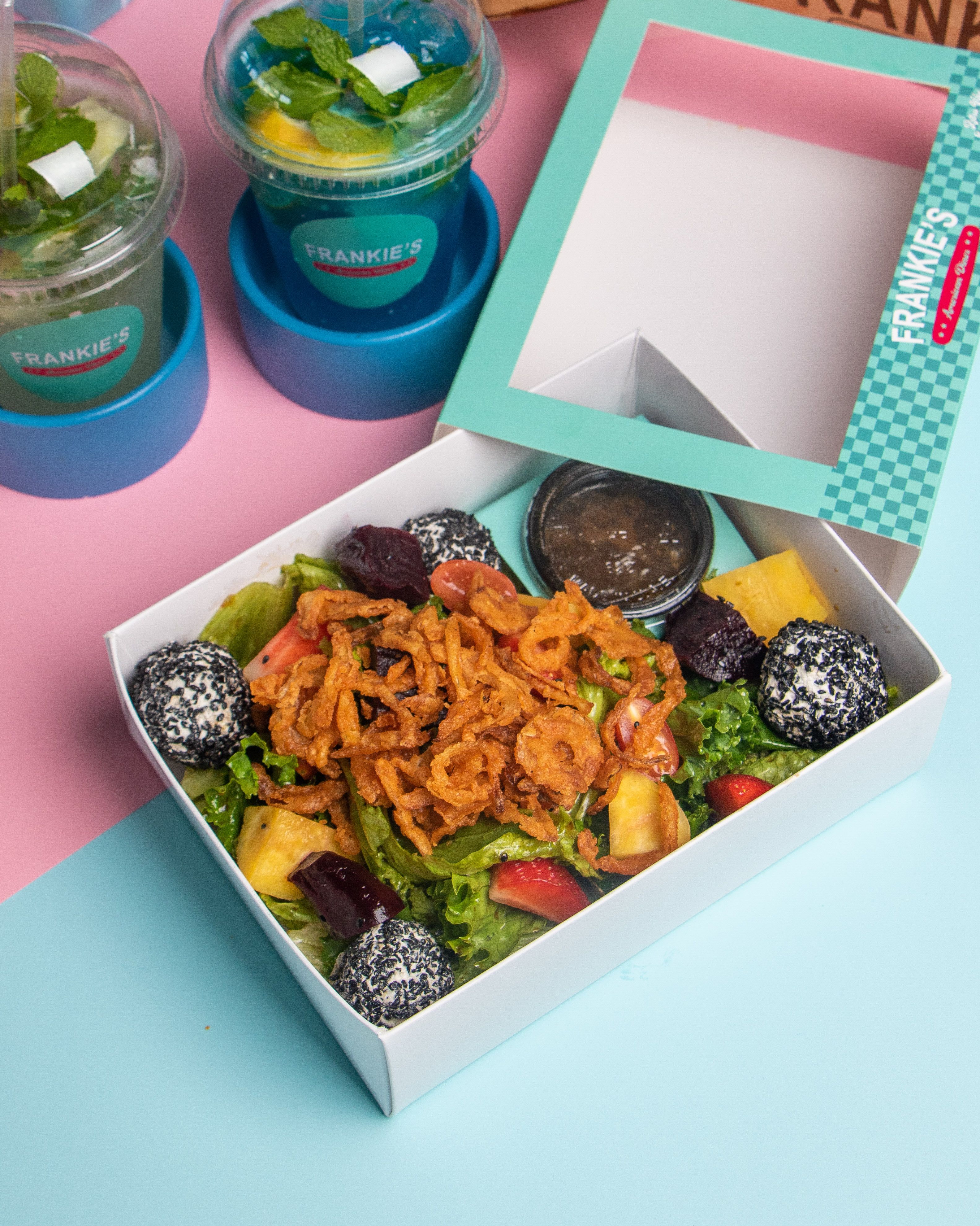 Colorful fresh salad in an open takeaway box with sauces and drinks on a pastel background.