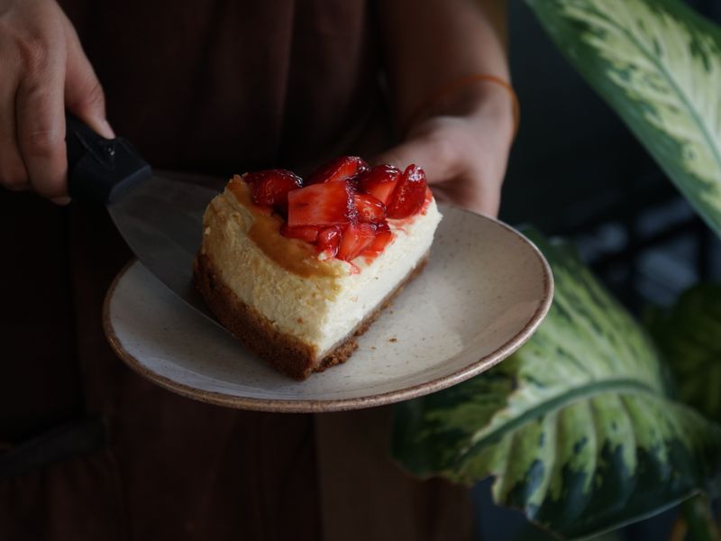 A delicious slice of strawberry-topped cheesecake served on a ceramic plate.