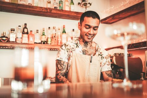 Free Tattooed bartender in apron preparing cocktails at a contemporary bar setting, surrounded by various liquor bottles. Stock Photo