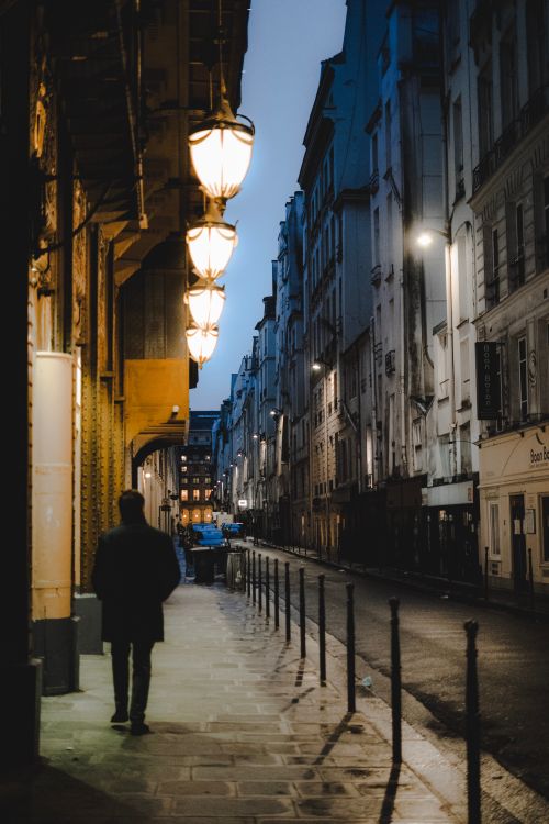Free A man walks alone on an urban street at dusk, with streetlights illuminating the cityscape in a serene atmosphere. Stock Photo