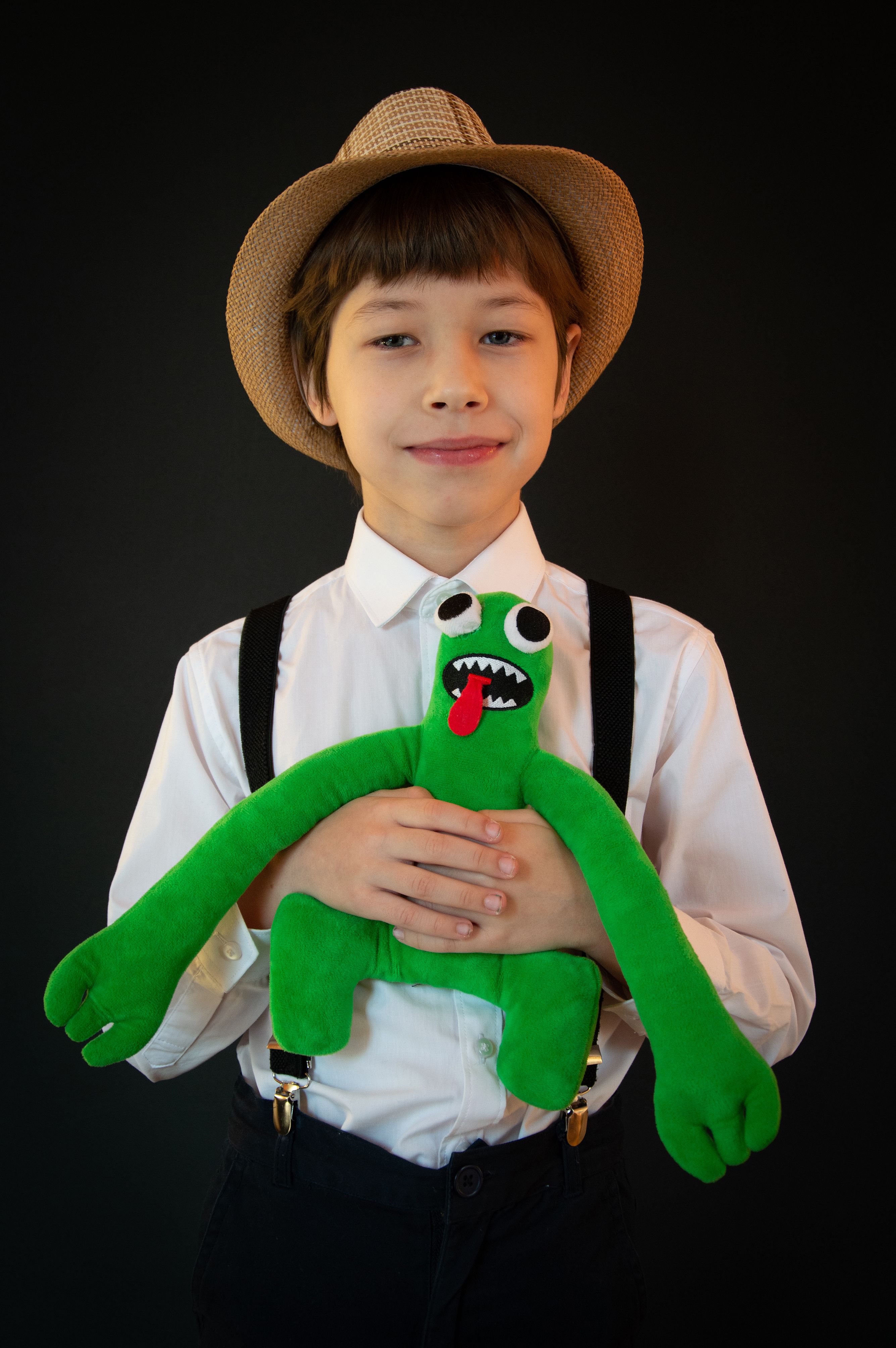 Charming portrait of a boy wearing a stylish hat and white shirt, holding a green plush toy, captured in a studio setting.