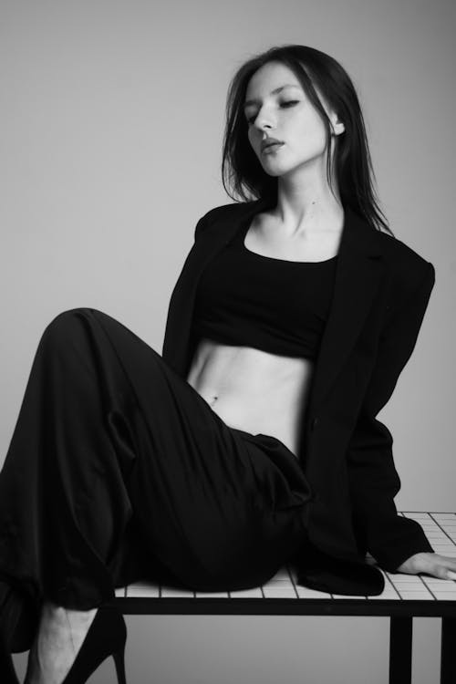 Grayscale Photography of a Woman in Blazer and Pants Sitting on a Table while Looking Afar