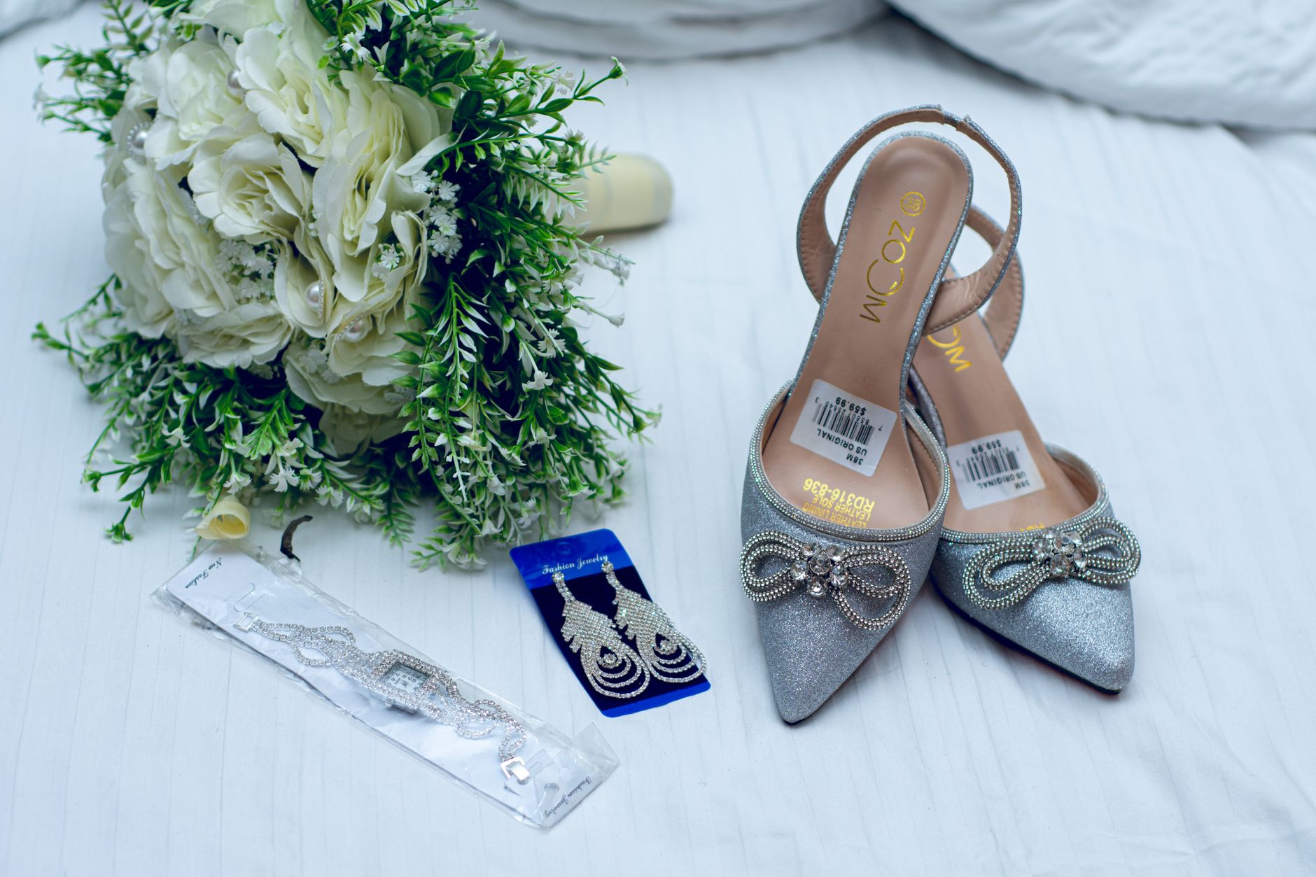 A close-up of wedding accessories including silver shoes, bouquet, and jewelry on a white surface.