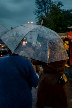 Two people walk in the rain with a clear umbrella under moody evening skies.