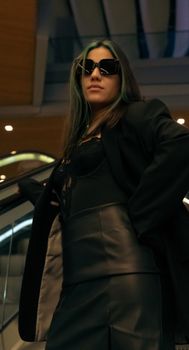 Fashionable adult woman in black leather outfit and sunglasses posing indoors.
