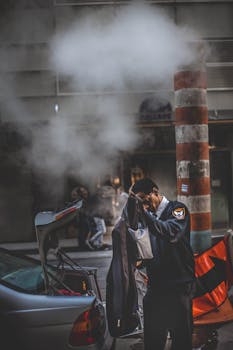 A security guard stands beside a car adjusting his coat on a steamy urban street.