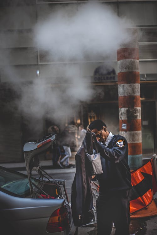 Free A security guard stands beside a car adjusting his coat on a steamy urban street. Stock Photo