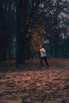 An adult man jogging through a park in Milan, Italy, surrounded by autumn leaves and trees.