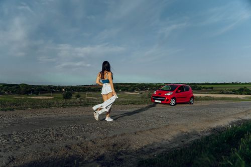 Free Young woman in denim walking on rural road towards a parked red car under a blue sky. Stock Photo