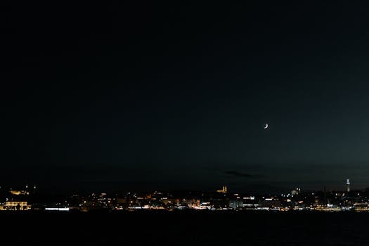 Urban skyline under a crescent moon, showcasing city lights at night.