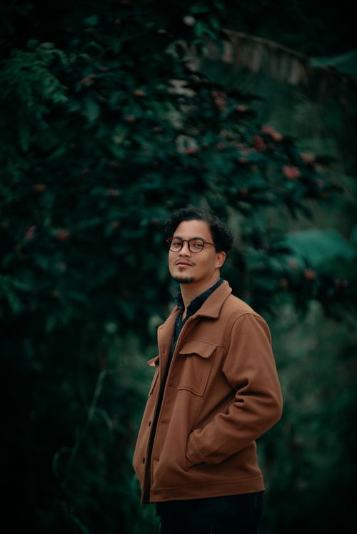 Free Stylish man in eyeglasses and brown jacket posing confidently outdoors with foliage background. Stock Photo