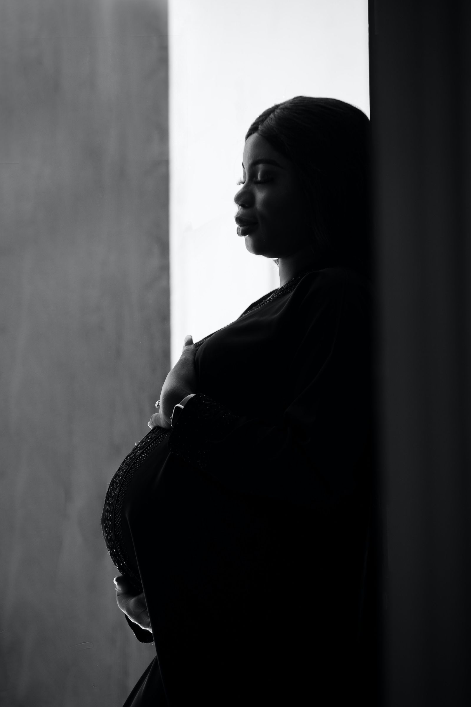 A pregnant Black woman leans against a wall in a black-and-white portrait, hand resting on her belly, contemplative expression.