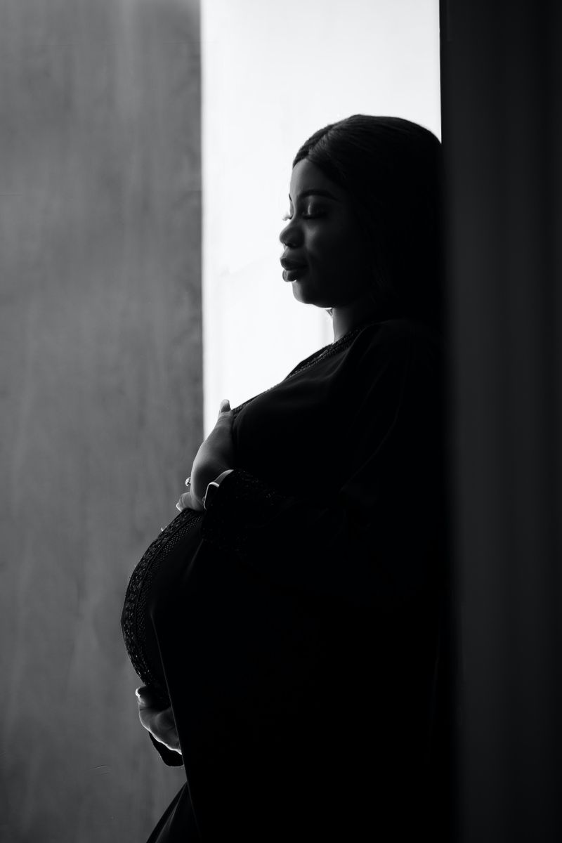 A pregnant Black woman leans against a wall in a black-and-white portrait, hand resting on her belly, contemplative expression.