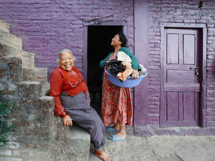 Elderly woman and daughter with laundry at vibrant purple house entrance.