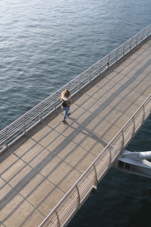 Free A woman walks on a seaside pier, casting shadows over the calm blue waters. Stock Photo