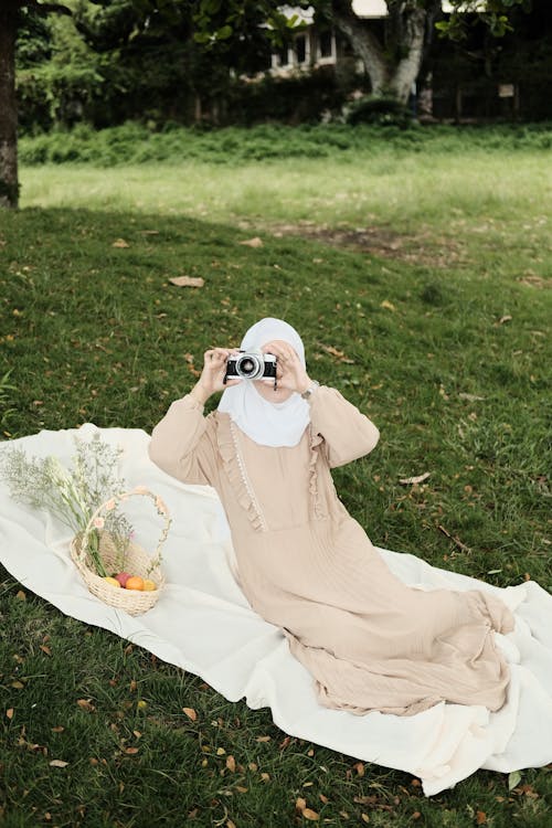 A Woman in White Hijab Sitting on a Picnic Blanket Taking Photos Using Camera