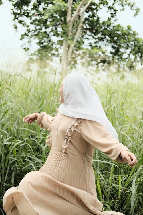 A Woman in White Hijab and Black Dress Standing on Grass Field while Spinning Around