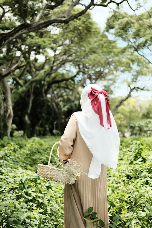Back View Shot of a Woman Wearing White Hijab with Red Ribbon and Brown Dress while Walking on Green Field