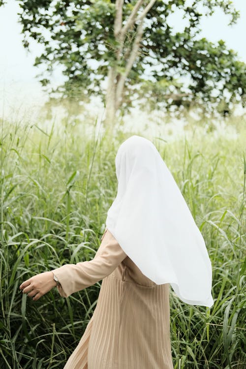 Back View Shot of a Woman Wearing White Hijab and Brown Dress Standing on Grass Field Near a Tree