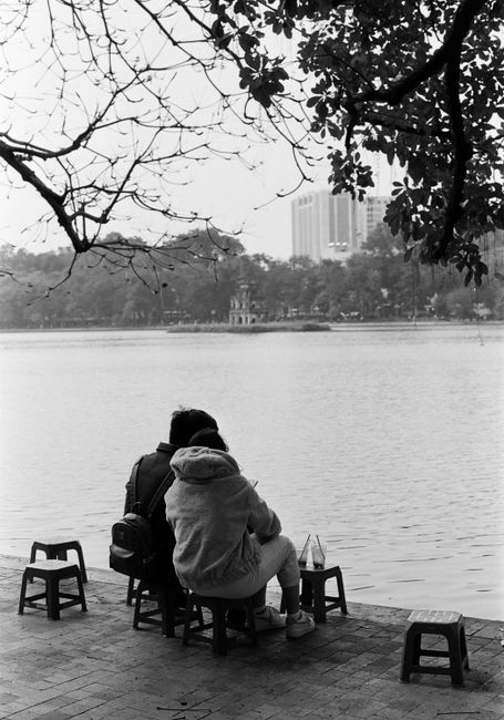 A couple sitting by a serene lake in a city, captured in black and white photography.