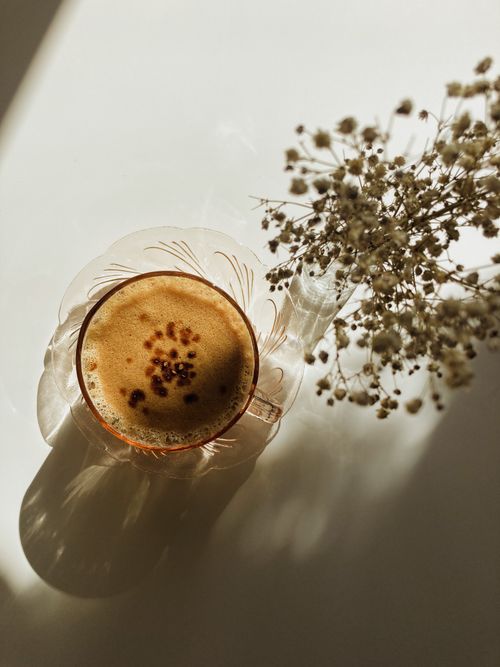 Free A delicate coffee in a glass cup beside a vase of flowers, casting artistic shadows. Stock Photo
