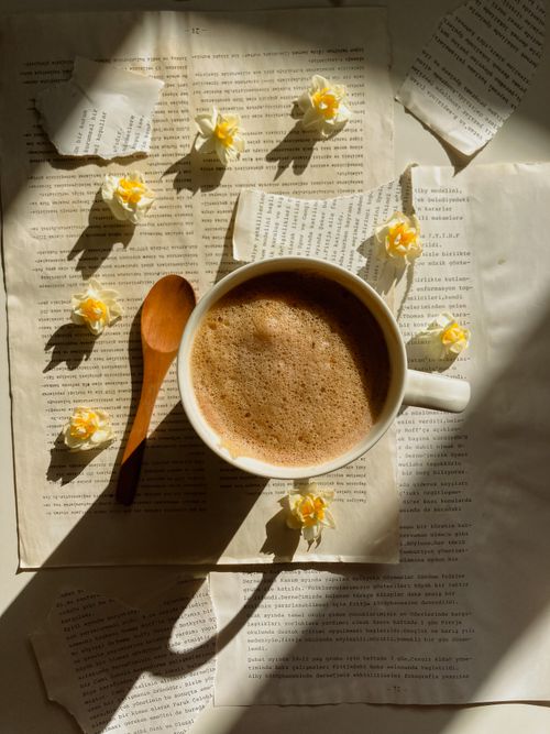 Free A cozy cup of frothy coffee on vintage texts, adorned with delicate yellow flowers and a wooden spoon. Stock Photo