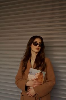 Elegant woman in sunglasses holding a magazine in front of textured background.