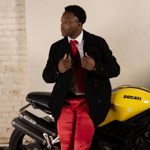Free A fashionable man in a red suit jacket poses confidently on a yellow Ducati motorcycle indoors. Stock Photo