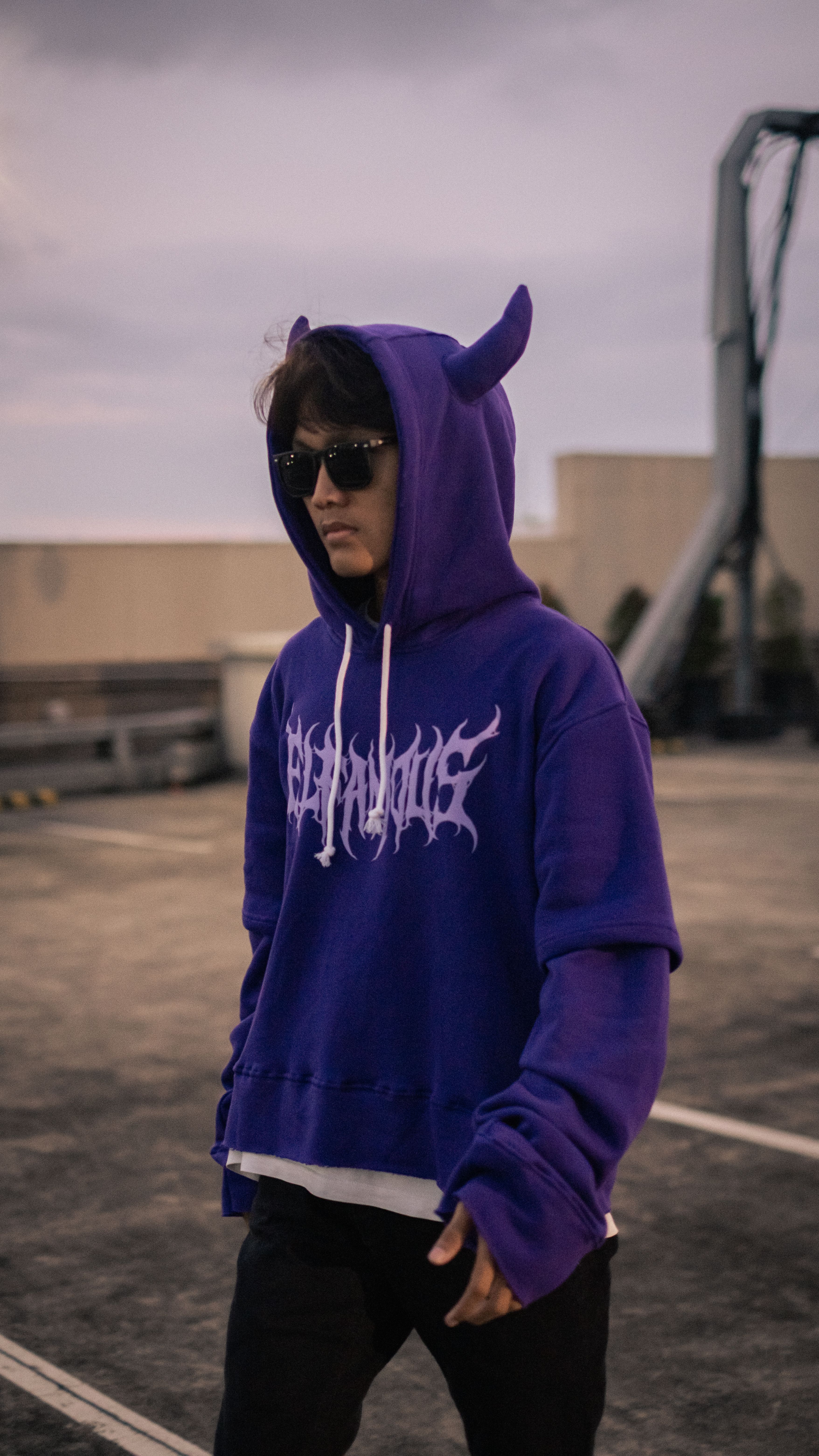A trendy young individual in a purple hoodie with horns, wearing sunglasses on a street.