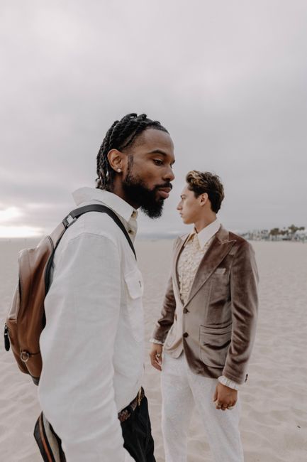 Two fashionable men in suits and casual attire on a cloudy beach day.