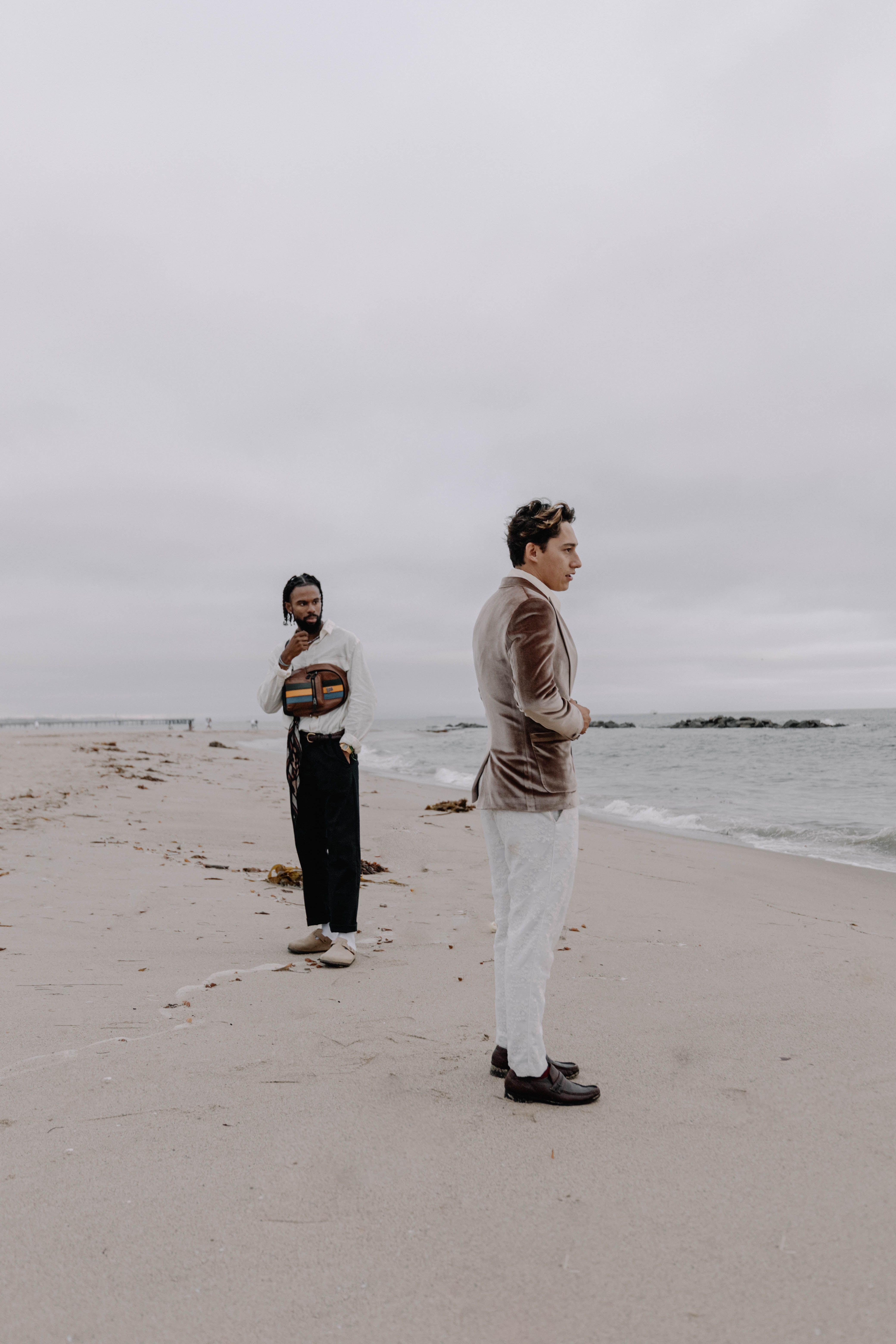 Stylish men in suits pose on a sandy beach under an overcast sky, creating a fashion-forward scene.