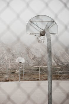 A misty basketball court with chain link fence and vintage hoops creates an atmospheric outdoor scene.