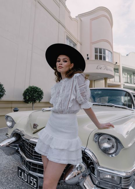 Elegant woman in white outfit and black hat leaning against a classic car in a stylish urban location.