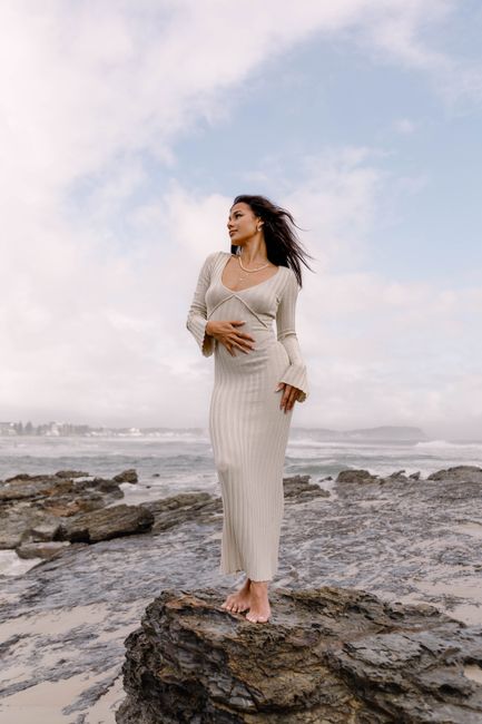 A woman in a stylish dress poses gracefully on a rocky beach with the sea and sky in the background.