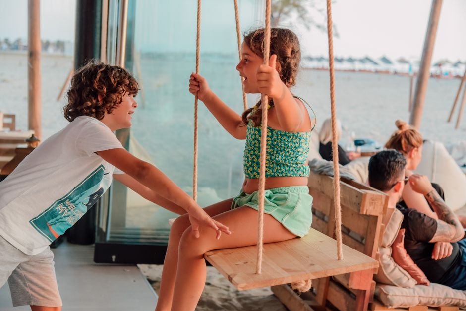 Niños disfrutando de un columpio en un café junto a la playa, capturando momentos de juego y la vibra del verano.