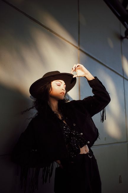 Fashionable woman in a hat posing against a shadowed wall with a stylish outfit.