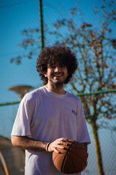 Young man with curly hair smiles while holding a basketball on an outdoor court.
