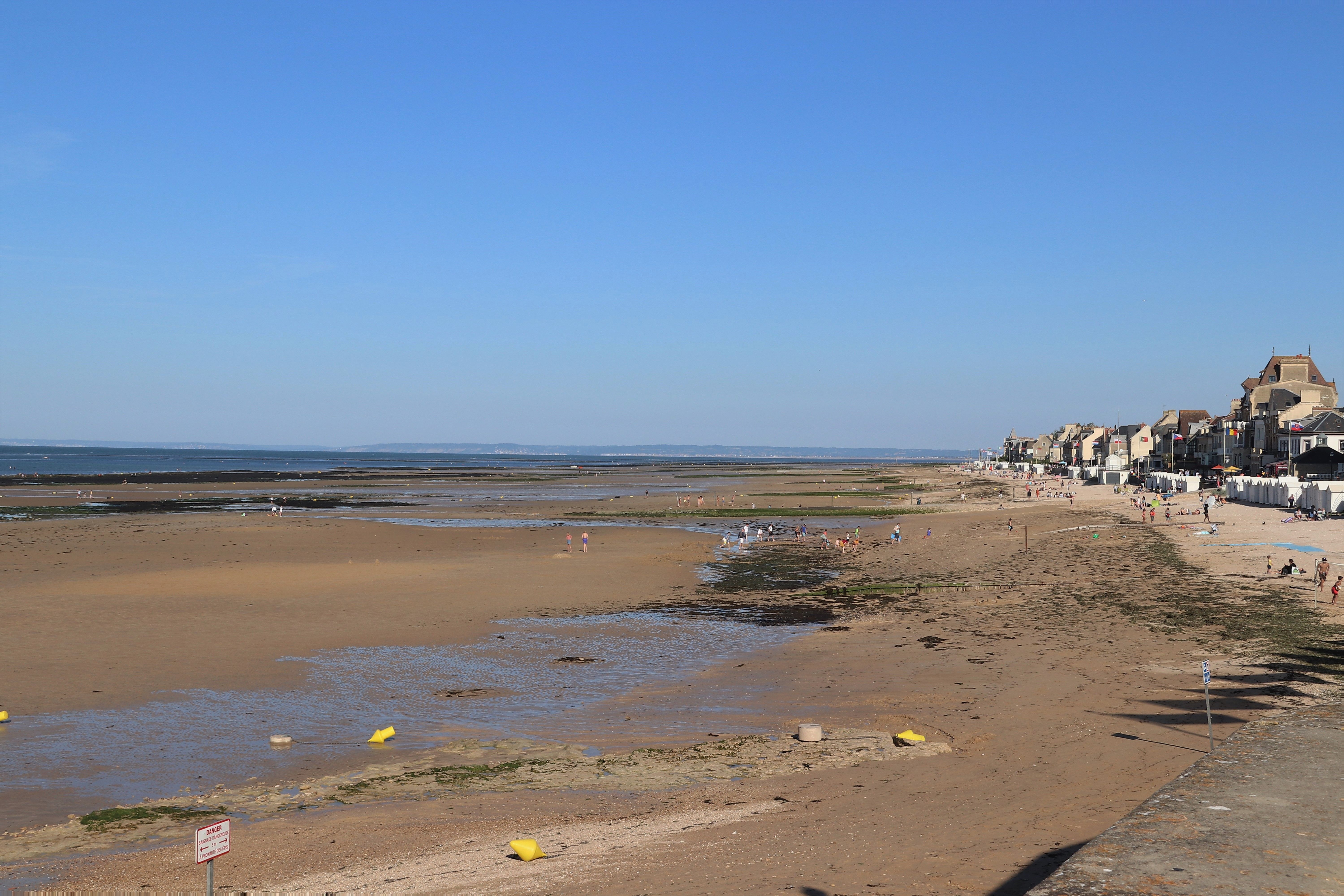 A sunlit beach view in Saint-Aubin-sur-Mer, Normandy, with coastal houses and low tide.