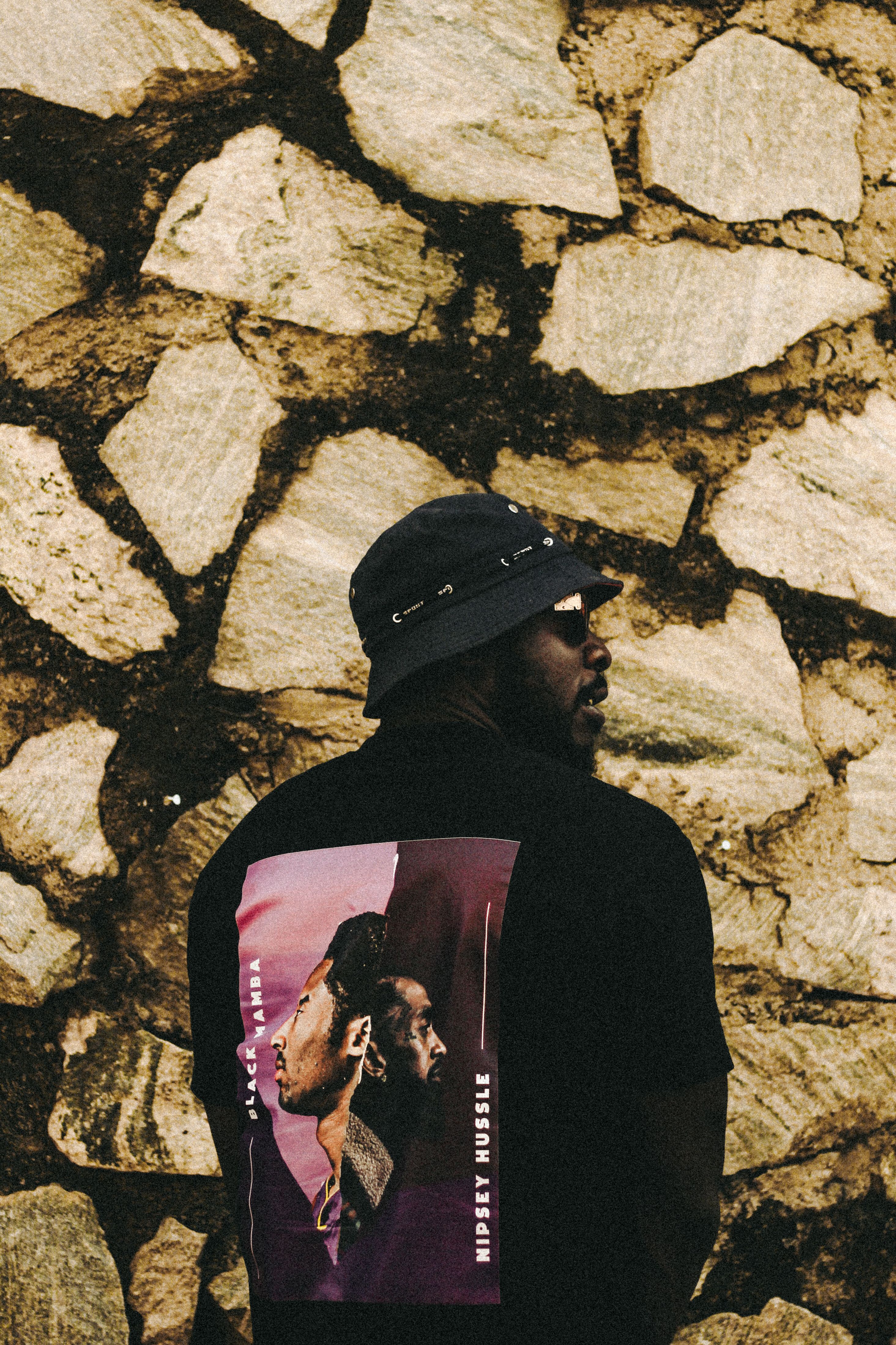 Stylish man in a bucket hat standing by a stone wall with artistic fashion elements.