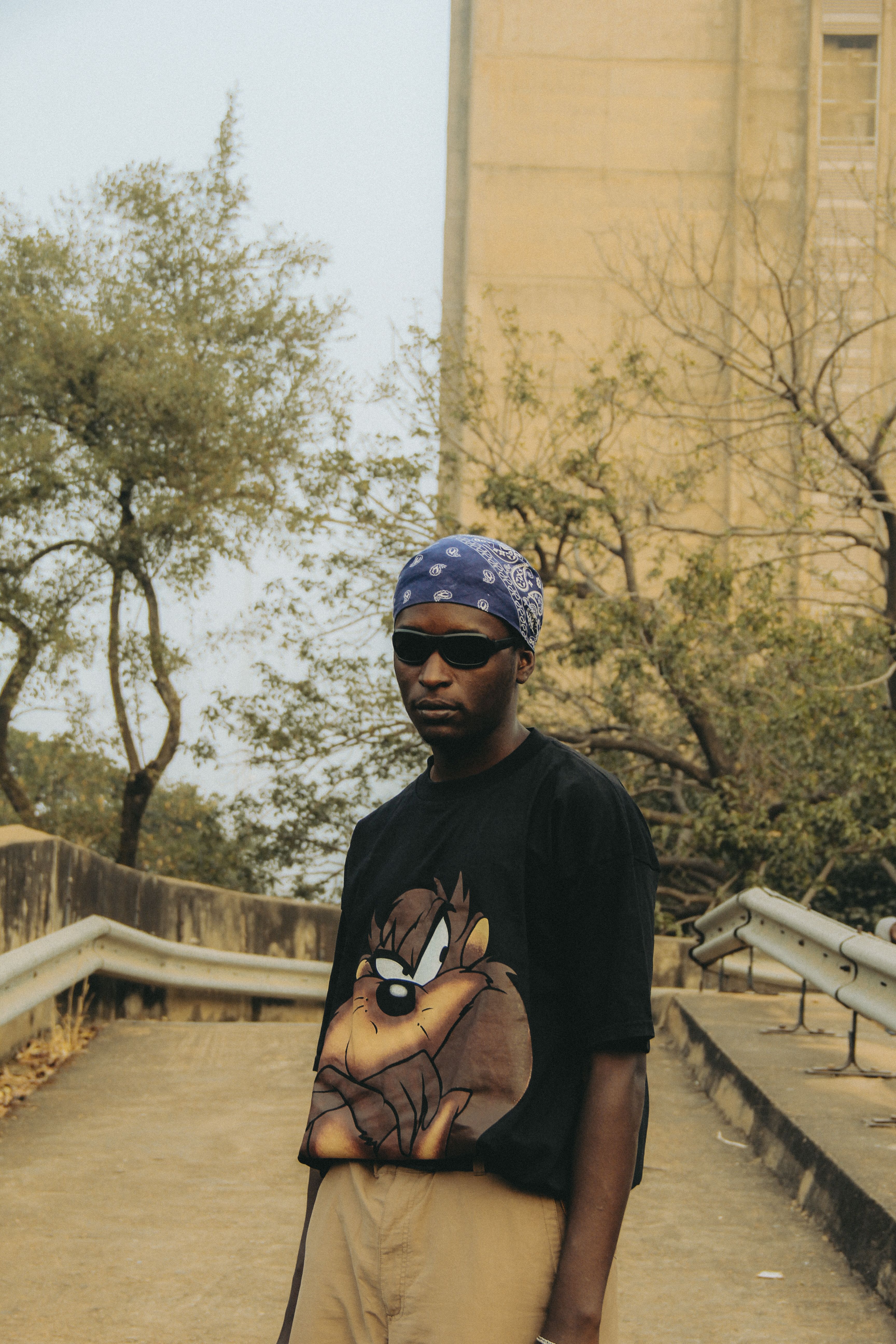 An African American man wearing sunglasses and a bandana stands stylishly outdoors in casual attire.