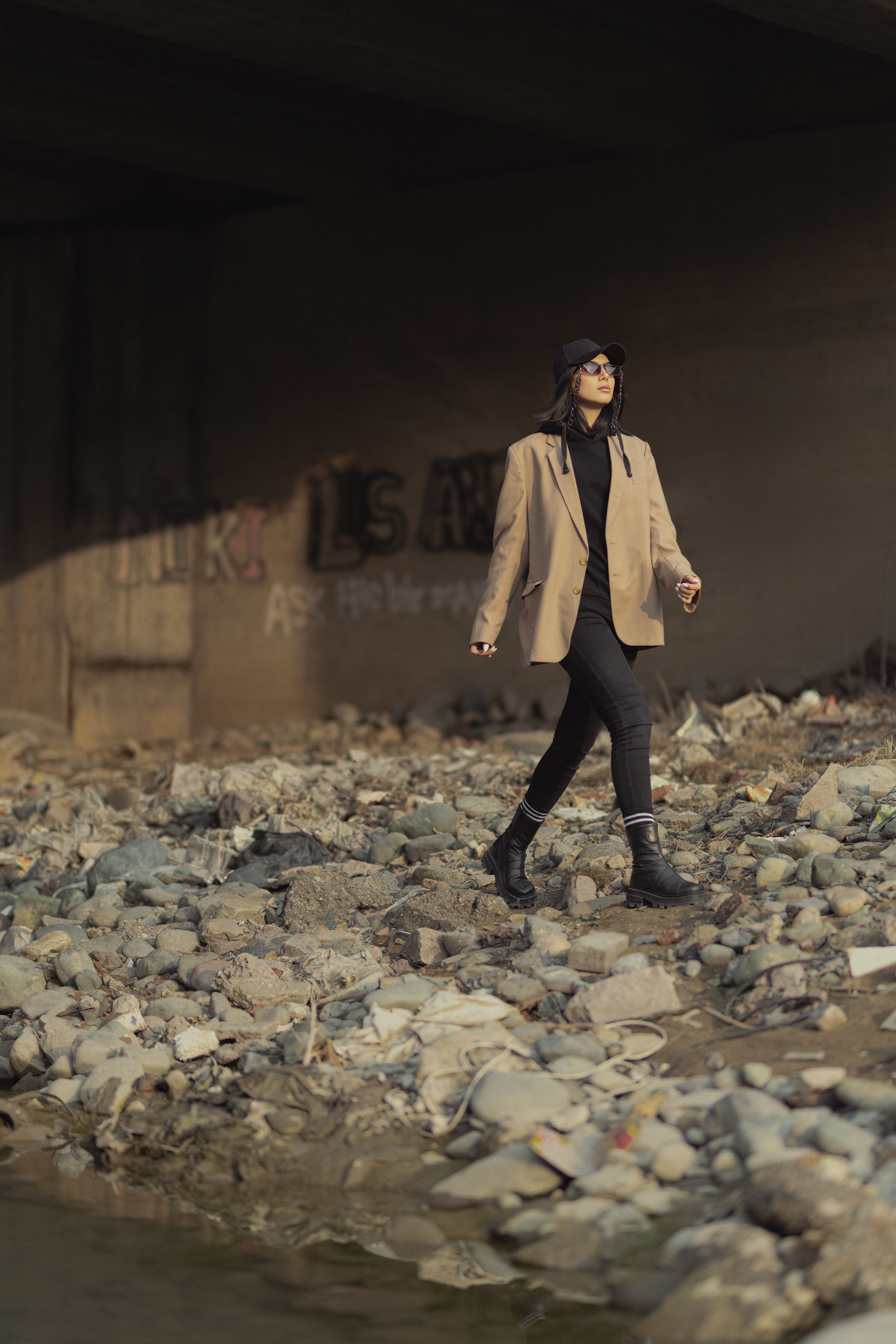 Stylish woman in a brown blazer and black boots walking over rocky ground under a bridge.