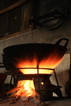 A large wok sits over a bright open fire against a rustic stone backdrop indoors.
