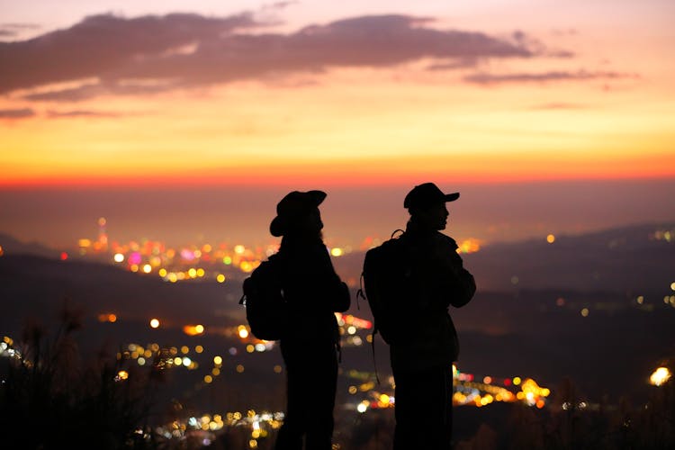 Silhouettes Of People Standing On Mountain Top Above City At Sunset