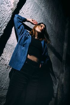 Stylish woman in a jean jacket and black top posing dramatically against a textured wall.