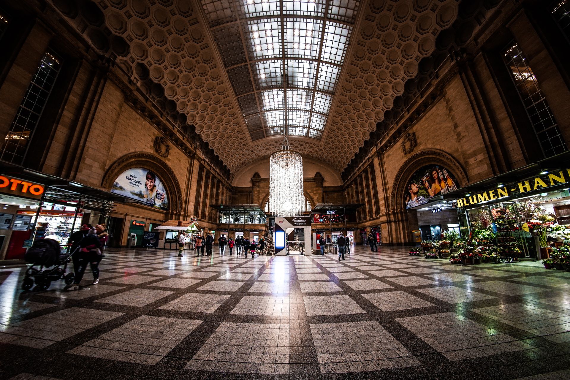 Bustling interior of Leipzig railway station with shops and travelers under ornate architecture.