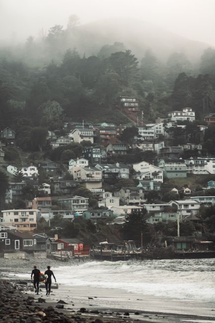 Two people walking along a misty beach with hillside houses in the distance.