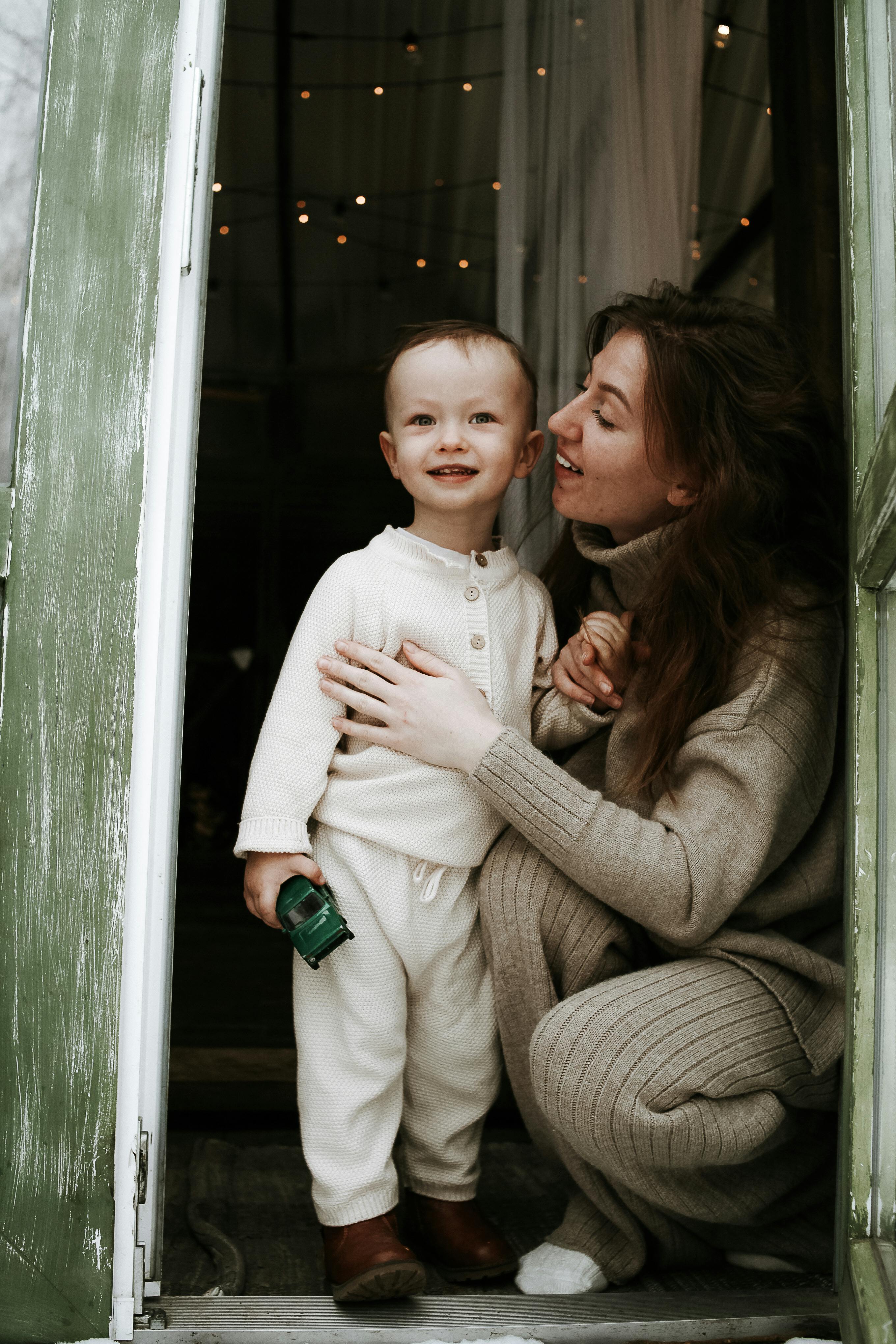 A happy mother crouching by her smiling child holding a toy in a cozy doorway scene.